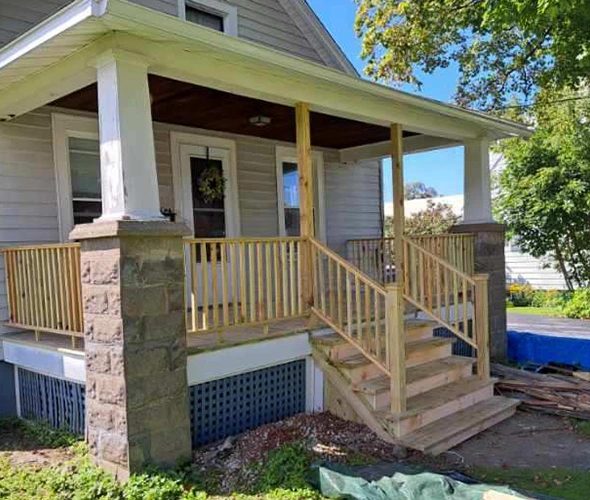 A construction worker carrying a large stack of lumber at a residential job site with a wooden house frame in the background.