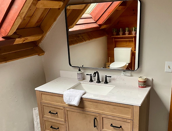 A modern bathroom remodel by Kennison Construction featuring a wood vanity with a white marble countertop, black hardware, and a large mirror reflecting a sloped wooden ceiling with a skylight.